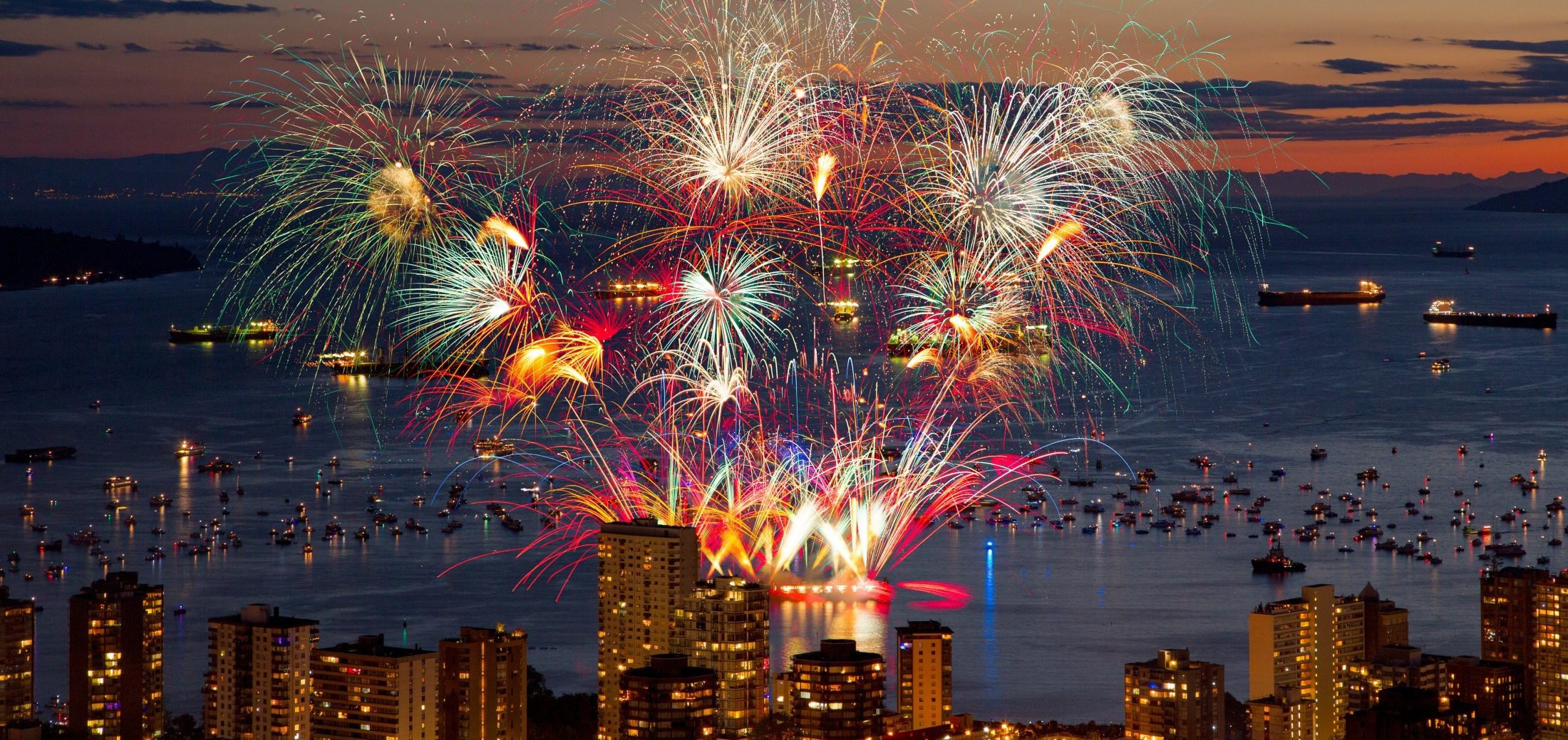 Fireworks in the sky above a body of water and buildings in the foreground.