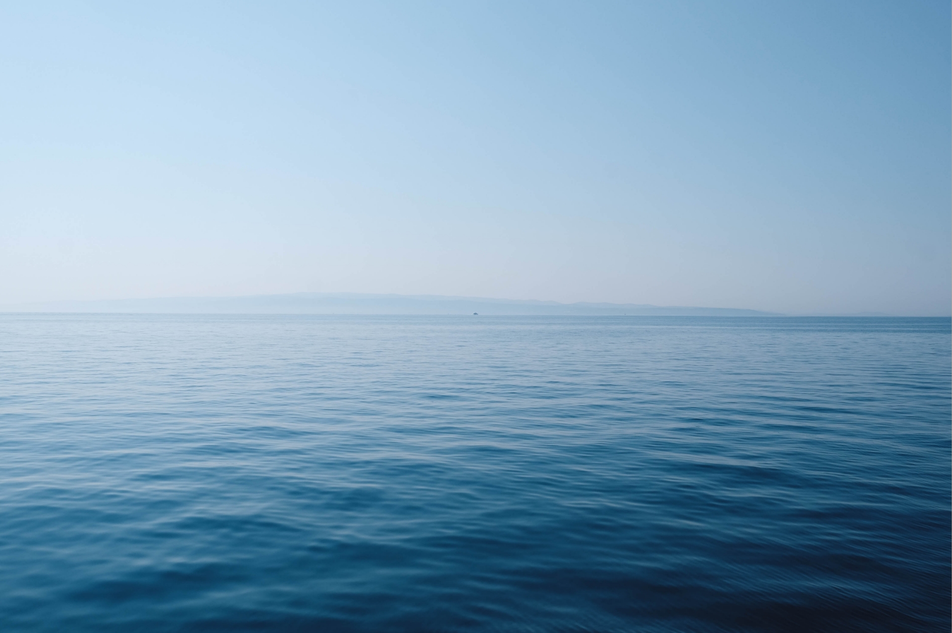A peaceful image of a calm, blue ocean stretching to the horizon under a clear sky, with distant land barely visible.
