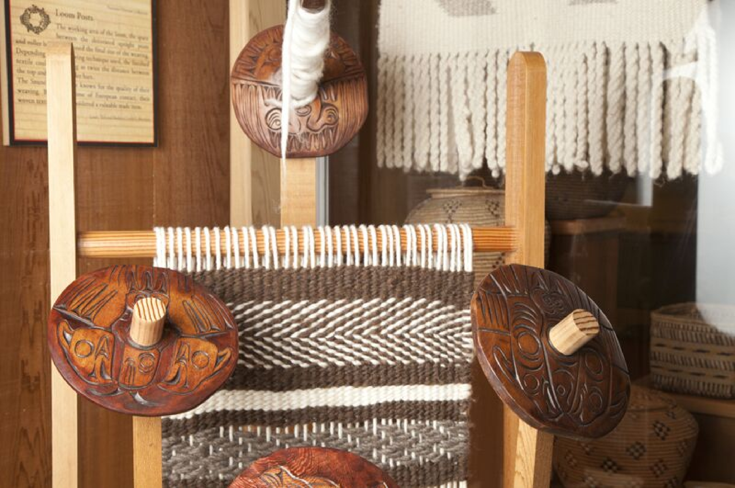 A close-up of a traditional weaving loom featuring intricate wooden carvings. The loom appears to be in a display setting, showcasing Indigenous designs in the wooden components and the woven textile in progress, with a mix of brown and cream threads.