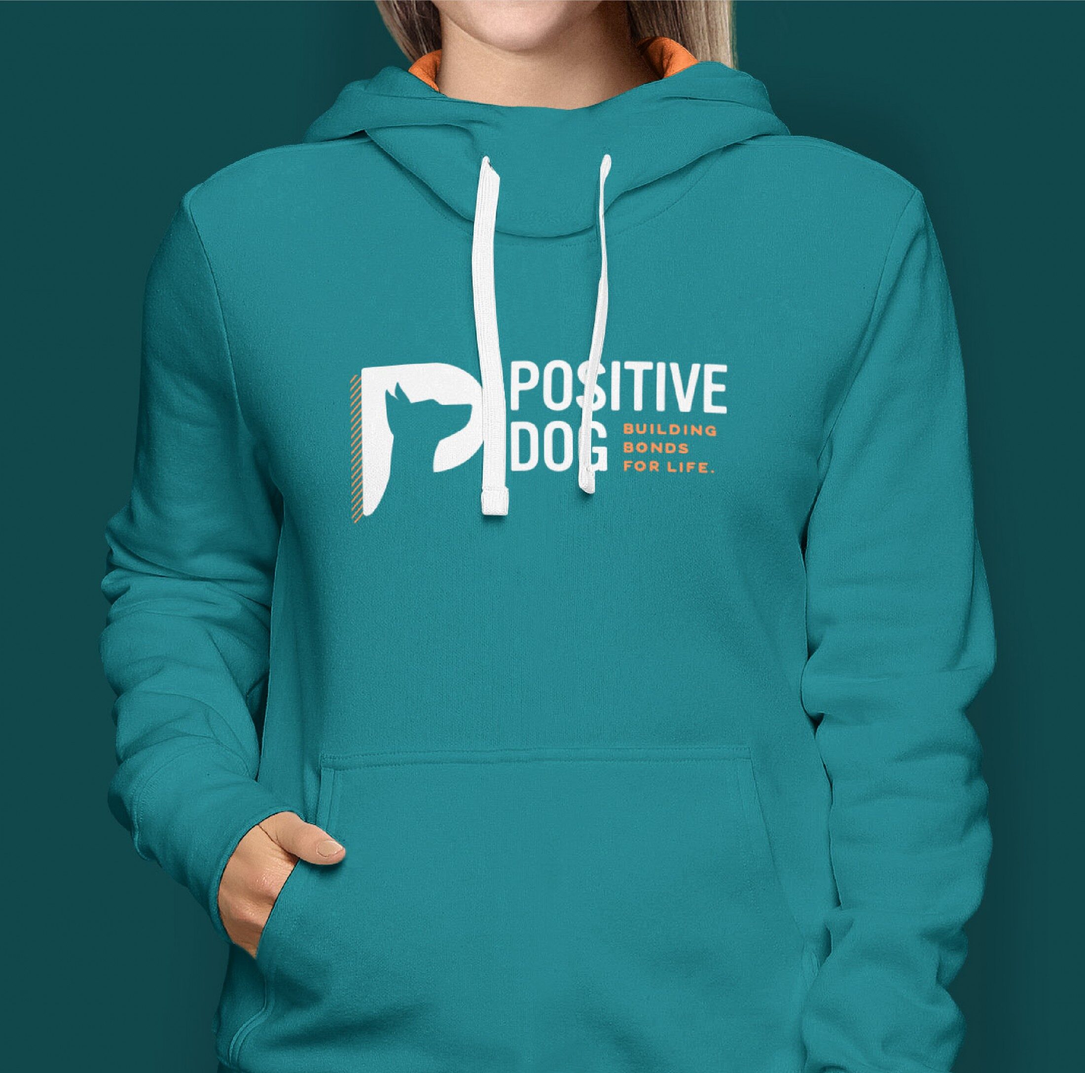 A person wearing a teal hoodie featuring the "Positive Dog" logo on the front. The logo includes the dog silhouette and the tagline "Building Bonds for Life," printed in white and orange.