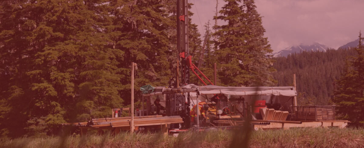 A drilling rig set up in a remote forested area with a background of dense trees and snow-covered mountain peaks in the distance.